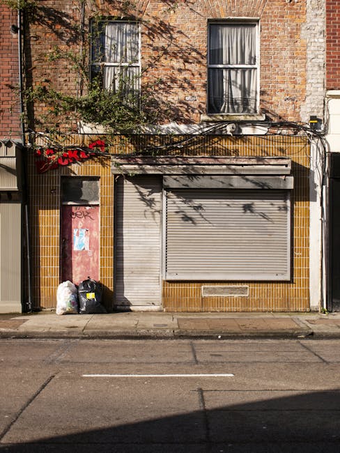 The image depicts the facade of a small commercial or residential building with a brick upper level and a ground floor clad in brown and yellow tiles. Two large, rectangular windows with white framing are situated on the upper level; the left window has partially drawn curtains, and the right window is also covered with curtains. The ground floor features a closed, grey metal roller shutter and an adjacent smaller access door, both situated within a tiled wall. In front of the building, on the pavement, there are three black garbage bags filled with waste, leaning against the left side of the entrance, with some white plastic bags beside them. Overhead, a network of black cables runs horizontally across the building's facade, casting shadows on the brickwork. The scene suggests a typical urban setting where private waste collection, possibly managed by House Clearance Stockwell, might be necessary for rubbish removal or on-site clearance, highlighting the need for professional waste management services in the area.