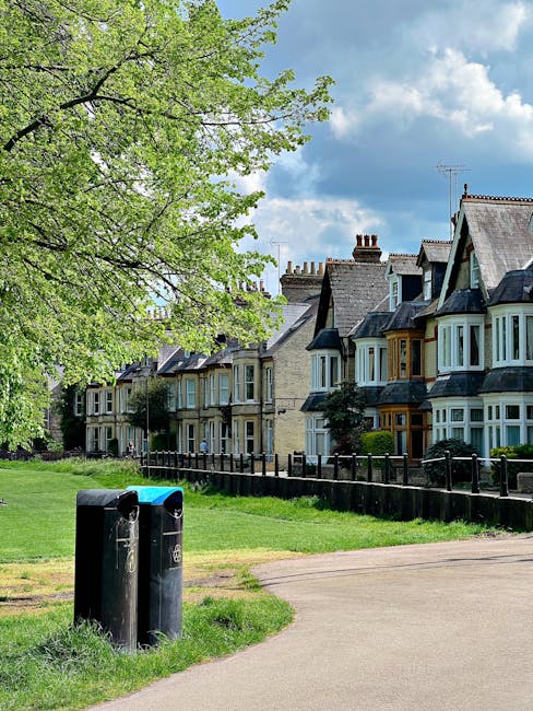 A row of traditional terraced houses with varying architectural features, including bay windows and gabled roofs, situated along a residential street. The houses have brick facades painted in neutral tones, with some displaying contrasting window frames in white and dark colors. In the foreground, there are two black waste bins, positioned near a grassy verge next to a paved footpath. A large, leafy green tree extends over part of the scene, casting dappled shade onto the grass and pathway, while a partly cloudy blue sky forms the backdrop. The scene reflects a typical suburban environment where private rubbish or waste collection services, such as those offered by House Clearance Stockwell, could be involved in local rubbish removal in the area, supporting the overall cleanliness and upkeep of residential neighbourhoods.