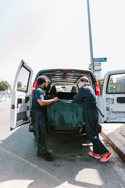 Two workers dressed in dark clothing and safety goggles are standing at the rear of a black van on a street, carefully loading a large, dark green upholstered piece of furniture into the vehicle's open cargo area. The van is parked on an asphalt road with a curb on the right side and a blue street sign visible above, indicating a residential or urban area. Bright sunlight illuminates the scene, casting shadows on the pavement. The interior of the van shows a spacious cargo area with walls and ceiling lined with black protective material, suitable for transporting bulky rubbish or household items. This scene presents a typical example of onsite rubbish removal or private waste handling, often undertaken when organising house clearances, aligning with services offered by companies like House Clearance Stockwell for alternative waste disposal solutions beyond local authority collection.