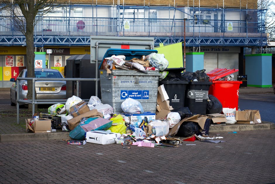 A large collection of mixed household waste and recyclable rubbish is piled on a paved sidewalk curbside in front of a commercial building, with multiple black, red, and grey plastic rubbish bins and open containers overflowing with paper, cardboard, plastic bags, and packaging materials. The waste includes flattened cardboard boxes, crumpled paper, plastic wrappers, and discarded packaging, some spilling onto the ground. Behind the rubbish, there is a parked silver car with a visible registration plate, and a metal railing separates the pile from the vehicle and sidewalk. The background features the facade of a building with a blue scaffolding structure, signage, and storefronts, indicating an urban retail or service area. The scene appears to be related to private rubbish disposal or on-site clearance services, which House Clearance Stockwell offers, and reflects the typical scenario of unscheduled or excess household waste awaiting collection in an urban environment.