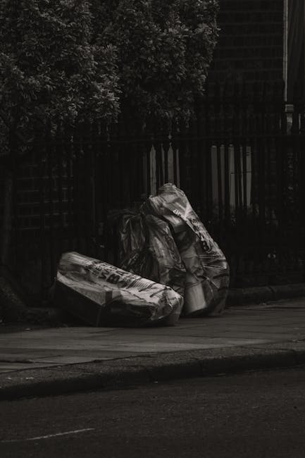 Two large black plastic rubbish bags, partially crumpled and tied at the top, are placed on a paved sidewalk adjacent to a street curb. The bags appear to contain mixed waste and are positioned near a black metal fence with vertical spindles, which runs along the edge of a residential or garden area. Behind the fence, dense foliage of evergreen shrubs or trees is visible, extending upward and partially obscuring a dark building structure with a staircase or balcony. The scene is captured in natural daylight, with shadows cast on the pavement, indicating a clear or partly cloudy day. The overall setting suggests a private property or an urban area where non-domestic waste is temporarily stored before collection. House Clearance Stockwell offers services for private waste removal, which is reflected in the available waste storage outside the property, ready for alternative rubbish collection or professional clearance.