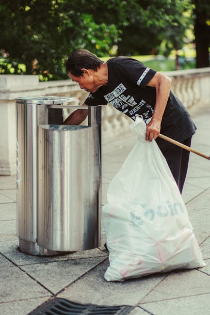 A large collection of mixed household waste and recyclable rubbish is piled on a paved sidewalk curbside in front of a commercial building, with multiple black, red, and grey plastic rubbish bins and open containers overflowing with paper, cardboard, plastic bags, and packaging materials. The waste includes flattened cardboard boxes, crumpled paper, plastic wrappers, and discarded packaging, some spilling onto the ground. Behind the rubbish, there is a parked silver car with a visible registration plate, and a metal railing separates the pile from the vehicle and sidewalk. The background features the facade of a building with a blue scaffolding structure, signage, and storefronts, indicating an urban retail or service area. The scene appears to be related to private rubbish disposal or on-site clearance services, which House Clearance Stockwell offers, and reflects the typical scenario of unscheduled or excess household waste awaiting collection in an urban environment.
